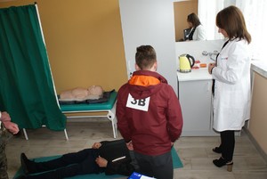 chłopak klęczy nad leżącym pokrzywdzonym w sali, gdzie odbywa się konkurencja udzielania pierwszej pomocy