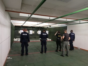 czterech policjantów na strzelnicy odwróconych plecami stoi w jednym rzędzie. Za nimi instruktor strzelania.