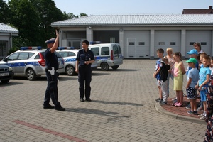 na placu parkingowym komendy policjant ruchu drogowego podnosi tarczę do zatrzymywania pojazdów. Wkoło grupa dzieci