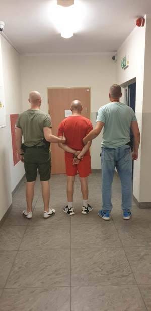 dwóch policjantów stojących tyłem na korytarzu pomieszczenia trzyma mężczyznę zakutego w kajdanki